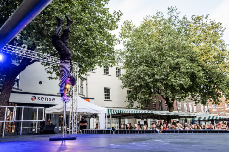 An acrobat is doing a hand stand on a metal apparatus on a stage outdoors. There is a crowd of people sat down below. The acrobat is dressed similar to a mime and is wear mime-like make up.